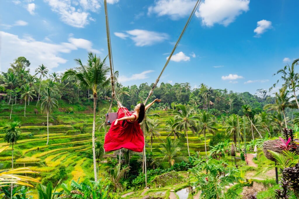 Happy young female tourist in red dress enjoying the Bali swing at tegalalang rice terrace in Bali